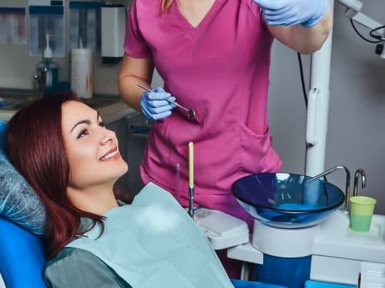 Periodontist in pink scrubs discussing gum disease treatment with smiling patient in dental chair.