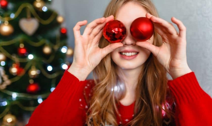 Young girl in red sweater holding red Christmas ornaments near her eyes, smiling with decorated tree behind her.