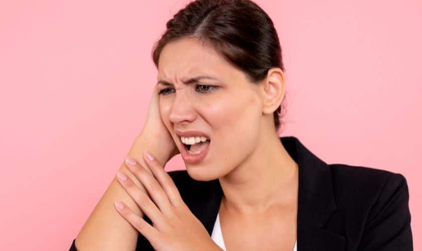 Woman experiencing tooth pain, holding her cheek in discomfort against a pink background.