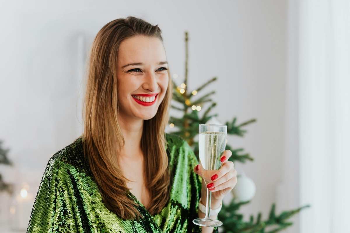 Surviving the Holiday Sugar Rush Oral Hygiene Tips for Christmas Treats Woman in green sequin top holding champagne glass near Christmas tree during holiday season.